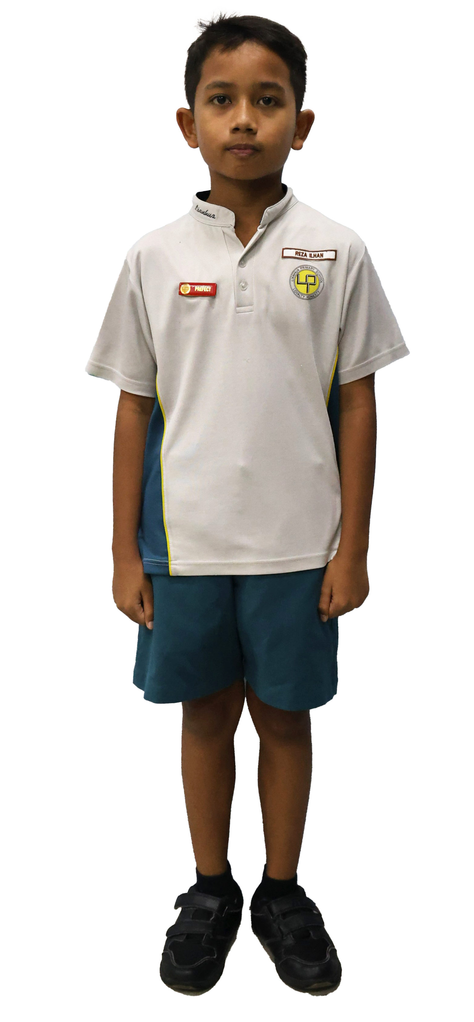 A young boy stands wearing a white school uniform shirt with badges, teal shorts, and black shoes.